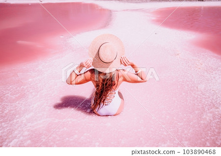 A woman traveler looks at an amazing pink salt lake. He sits with his back in a bathing suit and holds his hat in his hands. Wanderlust travel concept 103890468