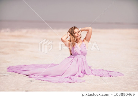 Pink lake woman. A woman in a pink dress sits on the salty shore of a pink lake and poses for a souvenir photo, creating lasting memories. 103890469