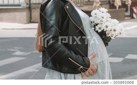 Close-up of a pregnant girl in a soft blue dress and black jacket with a bouquet of white chrysanthemums 103891008