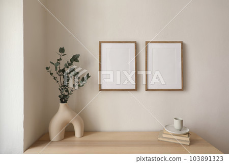 Two vertical picture frame mockup hanging on beige wall. Cup of coffee, books on wooden table. Vase with silver eucalyptus tree branches. Elegant interior, beautiful home decor. Trendy working space. 103891323