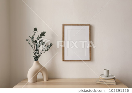 Empty vertical picture frame mockup hanging on beige wall. Cup of coffee, books on wooden table. Vase with silver eucalyptus tree branches. Minimal working space, home office. Scandinavian interior. 103891326