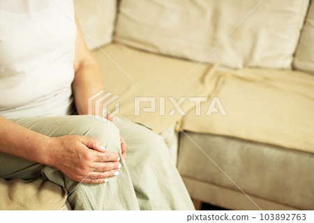 Senior woman suffering from knee pain on a sofa. People, health care and problem concept. Senior woman suffering from knee pain on a sofa. People, health care and problem concept. 103892763