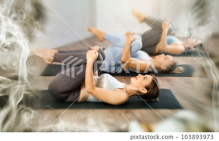 Diligent women practicing wind relieving pose of yoga in light fitness room Diligent women practicing wind relieving pose of yoga in light fitness room 103893973