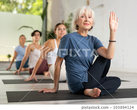 Relaxed positive old woman sitting in twisted position during group yoga training in studio Relaxed positive old woman sitting in twisted position during group yoga training in studio 103894124