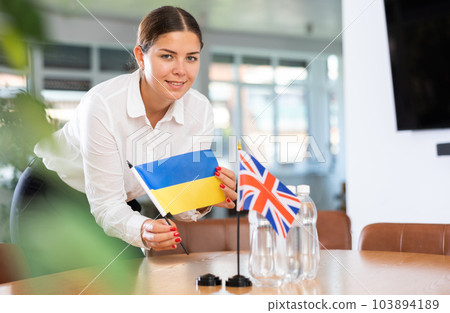 Positive young woman putting little flag of Ukraine on table next to the flag of Great Britain Positive young woman putting little flag of Ukraine on table next to the flag of Great Britain 103894189