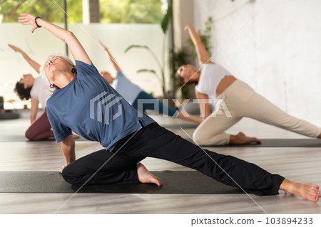 Old female stretching to sides while siting in Lotus pose during group yoga class in gym Old female stretching to sides while siting in Lotus pose during group yoga class in gym 103894233