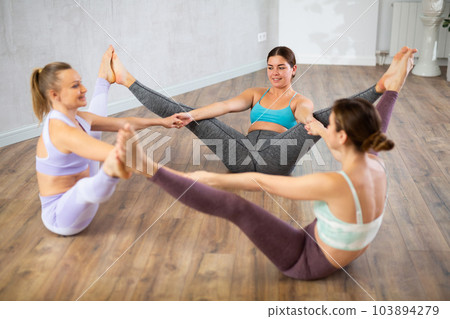 Smiling young women sitting on flour, holding hands and touching each others feet after training together in fitness center 103894279
