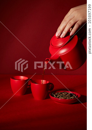 Female hand holding a red pot and pours tea in cup. Set of red pot and cup decorated on dark red background. Minimal abstract background for advertising product. Female hand holding a red pot and pours tea in cup. Set of red pot and cup decorated on dark red background. Minimal abstract background for advertising product. 103897199