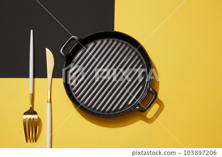 An empty black pan placed on black and yellow background with set of knife and fork. View from above. Rules of etiquette. Abstract minimal scene for advertising An empty black pan placed on black and yellow background with set of knife and fork. View from above. Rules of etiquette. Abstract minimal scene for advertising 103897206
