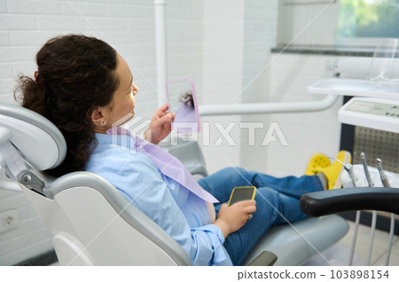Smiling pregnant woman, female patient sitting in dentist's chair, looking in the mirror after professional dental treatment in dentistry clinic. Dental practice. Caries prevention and teeth treatment Smiling pregnant woman, female patient sitting in dentist's chair, looking in the mirror after professional dental treatment in dentistry clinic. Dental practice. Caries prevention and teeth treatment 103898154