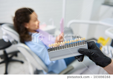 Vita scale. Bleaching teeth guide. Tooth color chart in the hands of a dentist orthodontist doctor, against blurred background of a female patient sitting in dental chair in modern dentistry clinic 103898168