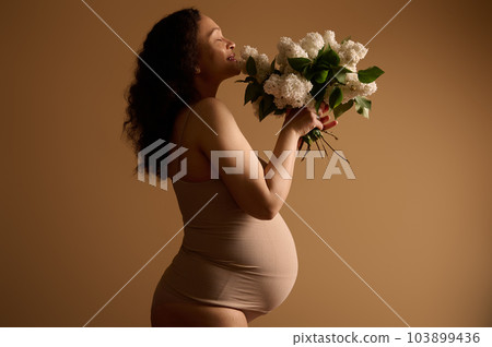 Side beauty portrait multi ethnic pretty pregnant gravid woman, expectant mother with big belly in pregnancy 28 week, wearing beige lingerie, holding white blooming lilacs, isolated studio background Side beauty portrait multi ethnic pretty pregnant gravid woman, expectant mother with big belly in pregnancy 28 week, wearing beige lingerie, holding white blooming lilacs, isolated studio background 103899436