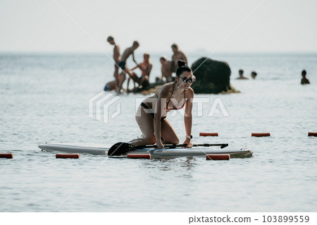 Sea woman sup. Silhouette of happy middle aged tanned woman in rainbow bikini, surfing on SUP board, confident paddling through water surface. Idyllic sunset. Active lifestyle at sea or river. 103899559