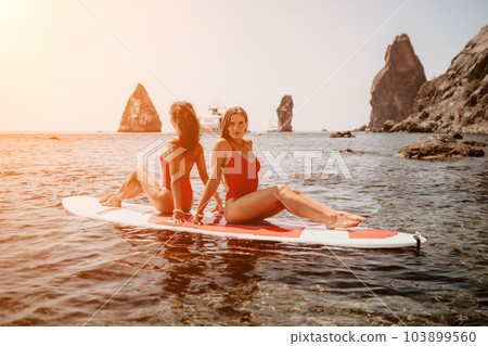 Woman sup yoga. Two happy sporty women practising yoga pilates on paddle sup surfboard. Female stretching doing workout on sea water. Modern individual female hipster outdoor summer sport activity. 103899560
