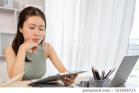 Beautiful young asian woman watching live video or video call of teacher teaching on laptop in her home, Take notes of important conversations and messages during the teacher's teaching. 103899723