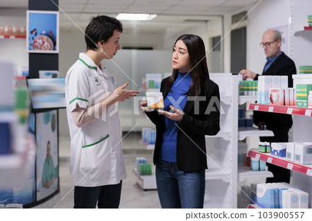 Asian woman holding vitamin package and listening to pharmacist explaining ascorbic acid dosage instruction. Drugstore customer buying food supplement, talking with pharmacy assistant in aisle Asian woman holding vitamin package and listening to pharmacist explaining ascorbic acid dosage instruction. Drugstore customer buying food supplement, talking with pharmacy assistant in aisle 103900557