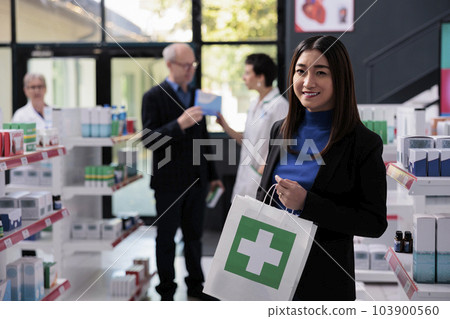 Smiling asian woman buying medication in drugstore and holding purchase portrait. Pharmacy store young buyer standing near shelf with medicament and carrying shopping paper bag Smiling asian woman buying medication in drugstore and holding purchase portrait. Pharmacy store young buyer standing near shelf with medicament and carrying shopping paper bag 103900560