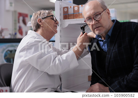 Old man getting ear examination procedure in pharmacy store, checking for elderly hearing loss problem. Woman pharmacist examining senior customer with otoscope medical tool in apothecary Old man getting ear examination procedure in pharmacy store, checking for elderly hearing loss problem. Woman pharmacist examining senior customer with otoscope medical tool in apothecary 103900582