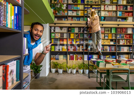 Young dark-skinned man hiding behind the bookshelves Young dark-skinned man hiding behind the bookshelves 103906013