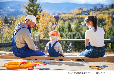 Back view of father, mother and son building wooden frame house. Toddler boy, daddy and woman having a rest on construction site. Guys wearing helmet and blue overalls. Carpentry, family concept. 103906929