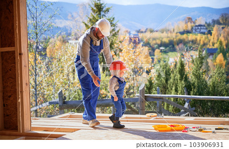 Father with toddler son building wooden frame house. Male builder playing with kid on construction site, wearing helmet and blue overalls on sunny day. Carpentry and family concept. 103906931