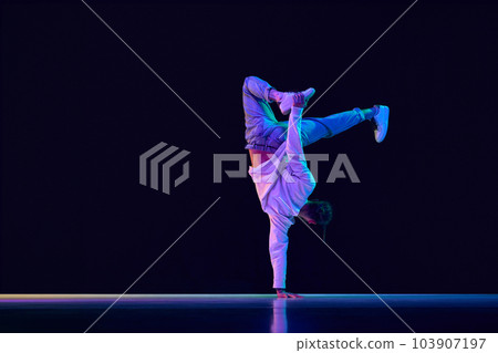Youth culture. Young man in casual clothes making performance, dancing breakdance against black studio background in neon light Youth culture. Young man in casual clothes making performance, dancing breakdance against black studio background in neon light 103907197