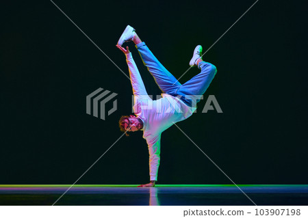 Dynamic image of talented, active young man in sport style clothes dancing breakdance against black studio background in neon light Dynamic image of talented, active young man in sport style clothes dancing breakdance against black studio background in neon light 103907198
