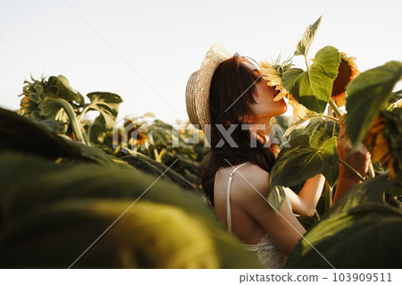 Smiling woman wearing a hat standing in a field of sunflowers in sunset Smiling woman wearing a hat standing in a field of sunflowers in sunset 103909511