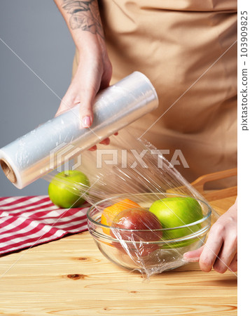 Woman putting cling filmover bowl of fresh fruit at wooden table Woman putting cling filmover bowl of fresh fruit at wooden table 103909825