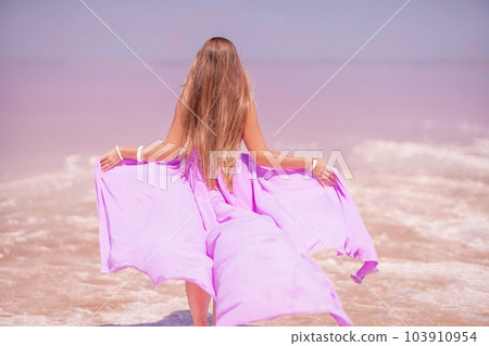 Woman pink salt lake. Against the backdrop of a pink salt lake, a woman in a long pink dress takes a leisurely stroll along the white, salty shore, capturing a wanderlust moment. 103910954