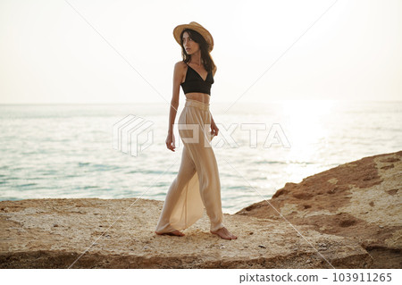 Young stylish woman standing on cliff at the sea at sunset Young stylish woman standing on cliff at the sea at sunset 103911265