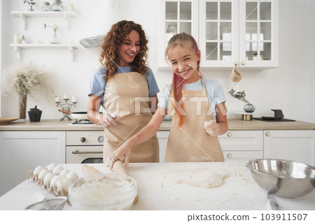 Teen girl helping her mom to cook dough in their kitchen at home Teen girl helping her mom to cook dough in their kitchen at home 103911507
