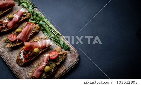 Bruschettas with jamon, olives, pesto, grilled cherry tomatoes served on wooden board with arugula on background with copy space. Mediterranean toasted bread with meat, cheese and vegetables 103912905