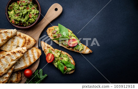 Bruschettas with toast, guacamole, basil and cherry tomatoes on wooden board served with arugula. Beautiful food composition with slice bread, avocado, herbs and vegetables with copyspace background 103912918