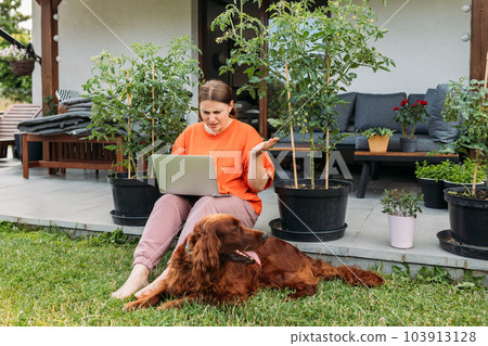 Beautiful Irish Setter dog is laying on grass, while girl working next to him. Photo of unhappy crazy angry woman scream laptop overworked bad mood sit in the yard of the house in summer. Full body 103913128