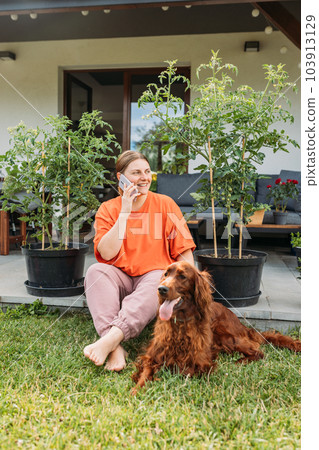 Beautiful Irish Setter dog is laying on grass, while girl using smartphone next to him. Cheerful happy young redhead woman relaxing with her dog in the yard of the house in summer. Full body 103913129