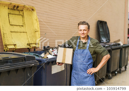 Garbage sorting with blond smiling man wearing blue apron. Man holds box filled with garbage near city trash can in back yard. Importance of responsible waste management and recycling practices 103914799