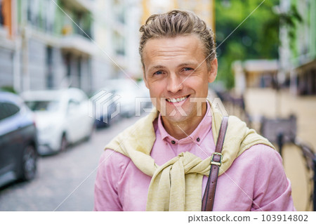 Handsome blond Caucasian man in an old city in Europe. With his charming smile and captivating blue eyes, he gazes directly at the camera, exuding confidence and positivity. High quality photo 103914802