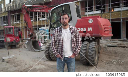 Portrait of successful builder, worker, contractor wearing hard hat and protective earphones, standing on work site with crossed arms confidently. Heavy construction equipment in background 103915985