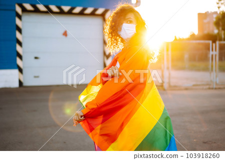 African American lesbian woman with medical face mask posing with lgbt pride flag. Curly woman during quarantine of coronavirus pandemic. African American lesbian woman with medical face mask posing with lgbt pride flag. Curly woman during quarantine of coronavirus pandemic. 103918260