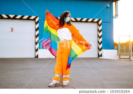 African American lesbian woman with medical face mask posing with lgbt pride flag. Curly woman during quarantine of coronavirus pandemic. 103918264