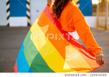 African American lesbian woman with medical face mask posing with lgbt pride flag. Curly woman during quarantine of coronavirus pandemic. African American lesbian woman with medical face mask posing with lgbt pride flag. Curly woman during quarantine of coronavirus pandemic. 103918269