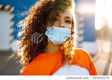 Young woman with medical face mask at sunset. Curly woman posing during quarantine of coronavirus pandemic. Young woman with medical face mask at sunset. Curly woman posing during quarantine of coronavirus pandemic. 103918270