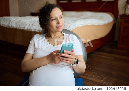 Multi-ethnic thoughtful pretty gravid female, pregnant woman, expectant mother holding mobile phone, checking new application, shopping online, dreamily looking aside, sitting on yoga mat at home Multi-ethnic thoughtful pretty gravid female, pregnant woman, expectant mother holding mobile phone, checking new application, shopping online, dreamily looking aside, sitting on yoga mat at home 103918636