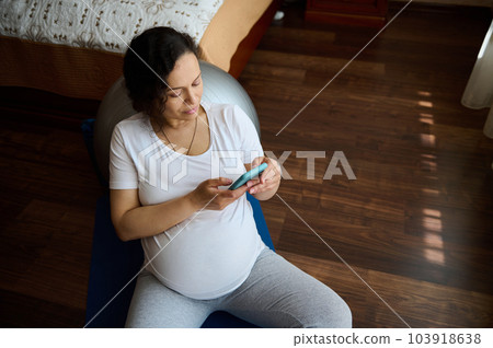 View from above of pensive beautiful adult pregnant woman using smart phone, checking mobile app, sharing maternity lifestyle on social media, sitting on floor in the bedroom. Happy carefree pregnancy View from above of pensive beautiful adult pregnant woman using smart phone, checking mobile app, sharing maternity lifestyle on social media, sitting on floor in the bedroom. Happy carefree pregnancy 103918638