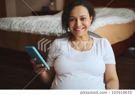 Close-up portrait of multi ethnic happy pregnant woman holding smartphone, smiling looking at camera. Pregnancy. People. Technology. Online shopping. Checking mobile application. Booking. Ordering Close-up portrait of multi ethnic happy pregnant woman holding smartphone, smiling looking at camera. Pregnancy. People. Technology. Online shopping. Checking mobile application. Booking. Ordering 103918639