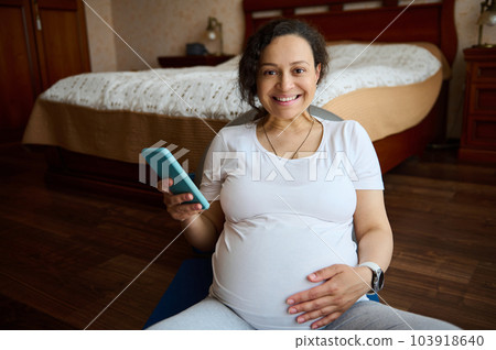 Portrait of a multi-ethnic smiling gravid woman, keeping her hand on her pregnant tummy, smiling looking at camera, relaxing on a yoga mat with a smartphone in hand. Happy pregnancy and maternity Portrait of a multi-ethnic smiling gravid woman, keeping her hand on her pregnant tummy, smiling looking at camera, relaxing on a yoga mat with a smartphone in hand. Happy pregnancy and maternity 103918640