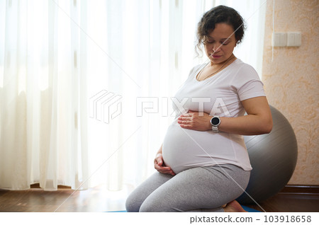 Confident expectant mother, gravid woman touching her pregnant belly, feeling baby kicks, sitting on a yoga mat in hero pose, against a window background, doing prenatal breathing exercises at home Confident expectant mother, gravid woman touching her pregnant belly, feeling baby kicks, sitting on a yoga mat in hero pose, against a window background, doing prenatal breathing exercises at home 103918658