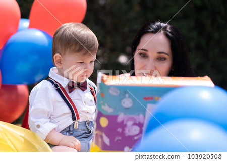 Exciting first birthday party in the open air - Little boy and mom holding wrapped present box, colorful balloons, and an unforgettable outdoor experience. Exciting first birthday party in the open air - Little boy and mom holding wrapped present box, colorful balloons, and an unforgettable outdoor experience. 103920508
