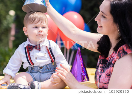 Festive outdoor gathering for a first birthday - Little boy and mom enjoy a lively party in the open air, balloons, creating a memorable first birthday celebration.  103920550
